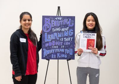 Two people standing with a poster in between them. The woman on the right-hand side is holding the book “Wisdom Tales”.