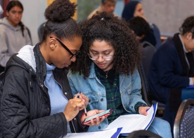 Two people holding papers sitting side-by-side looking at a phone.
