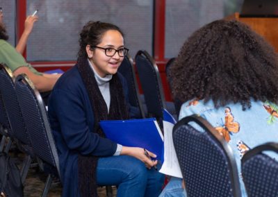 Two people sitting among a row of chairs turned to each other and are talking.