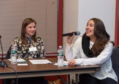 Two people sitting at a table. The person on the right-hand side is holding a microphone and speaking.