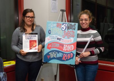 Two people standing with a poster in between them. The person on the left-hand side is holding the book “Wisdom Tales”.
