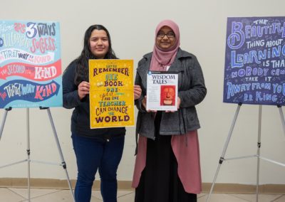 Two people standing side-by-side slightly in front of two posters that are on the left and right hand sides of the room. The person on the left-hand side is holding up a poster. The person on the right-hand side is holding up a book.