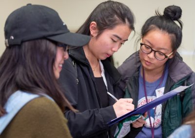 Three people standing together. The person in the middle is holding a paper and taking notes while others look on.