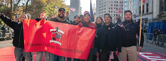 Students holding up a red banner that reads “Queens College Veterans Club”.