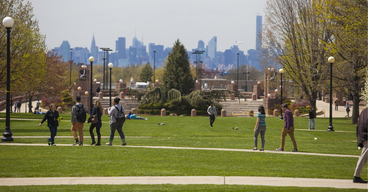 People walking through Queens College Campus with the New York City skyline visible in the background.
